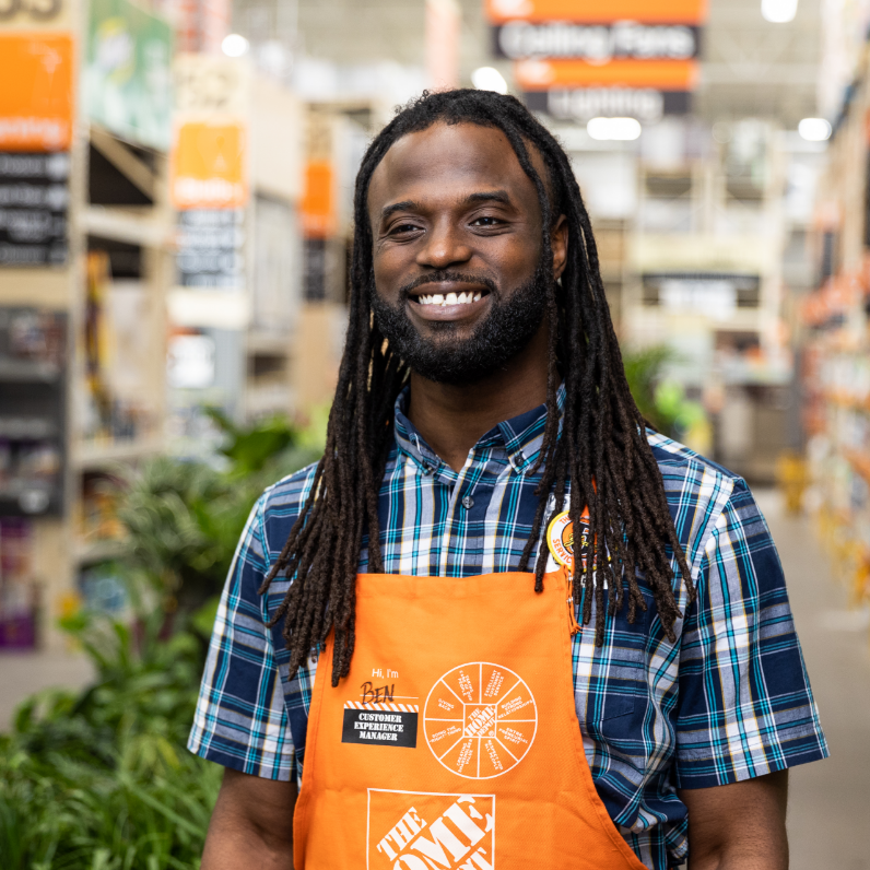 A young black man in a checkered polo and orange apron smiles to the camera while standing in an aisle.