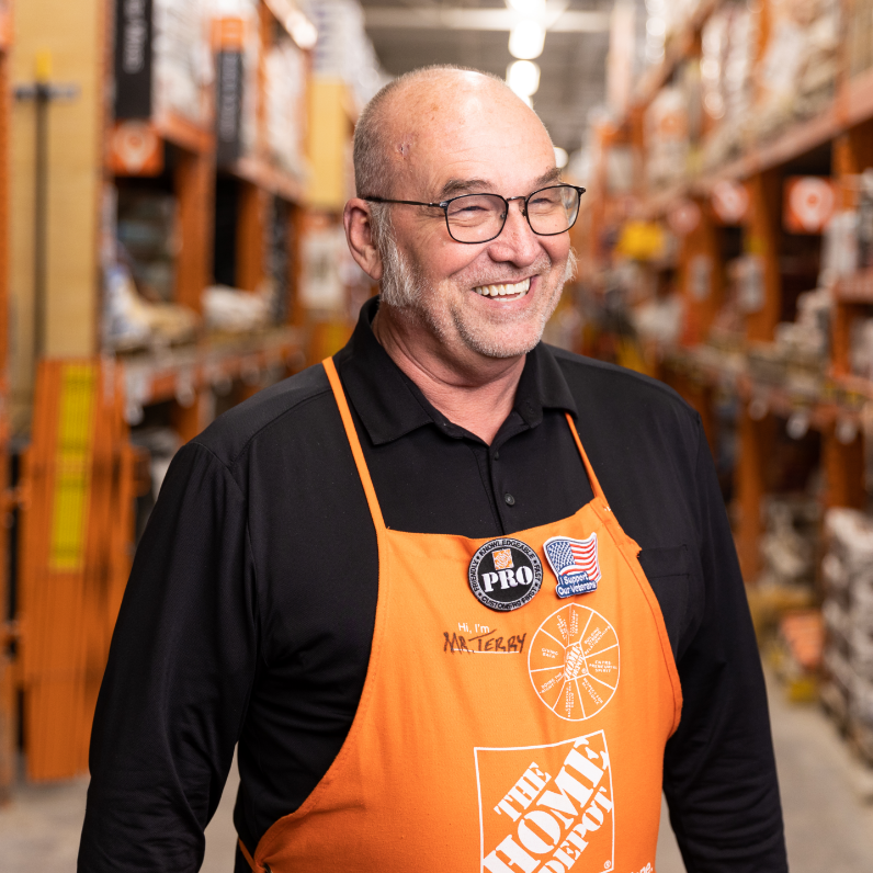 Elderly bald white man in black shirt and orange apron smiles to the right.