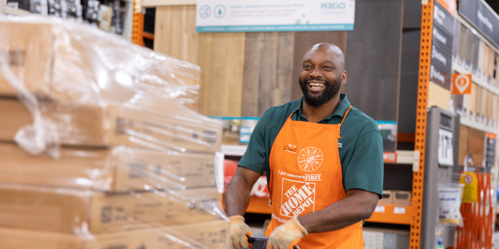 Middle-aged black man in a green polo and orange apron smiles as he transports boxes in a store.